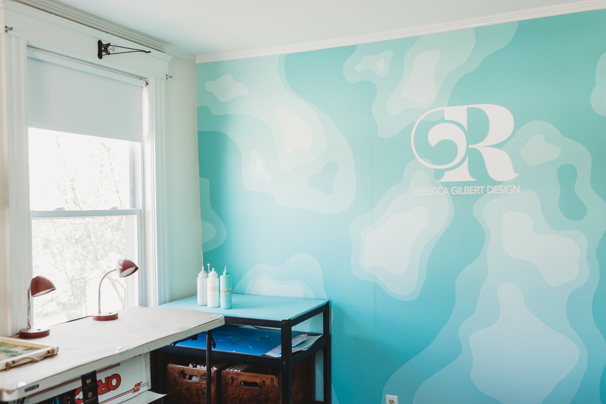 Room with a teal wall featuring a topographic map, rebecca gilbert design logo, a desk, and a window.
